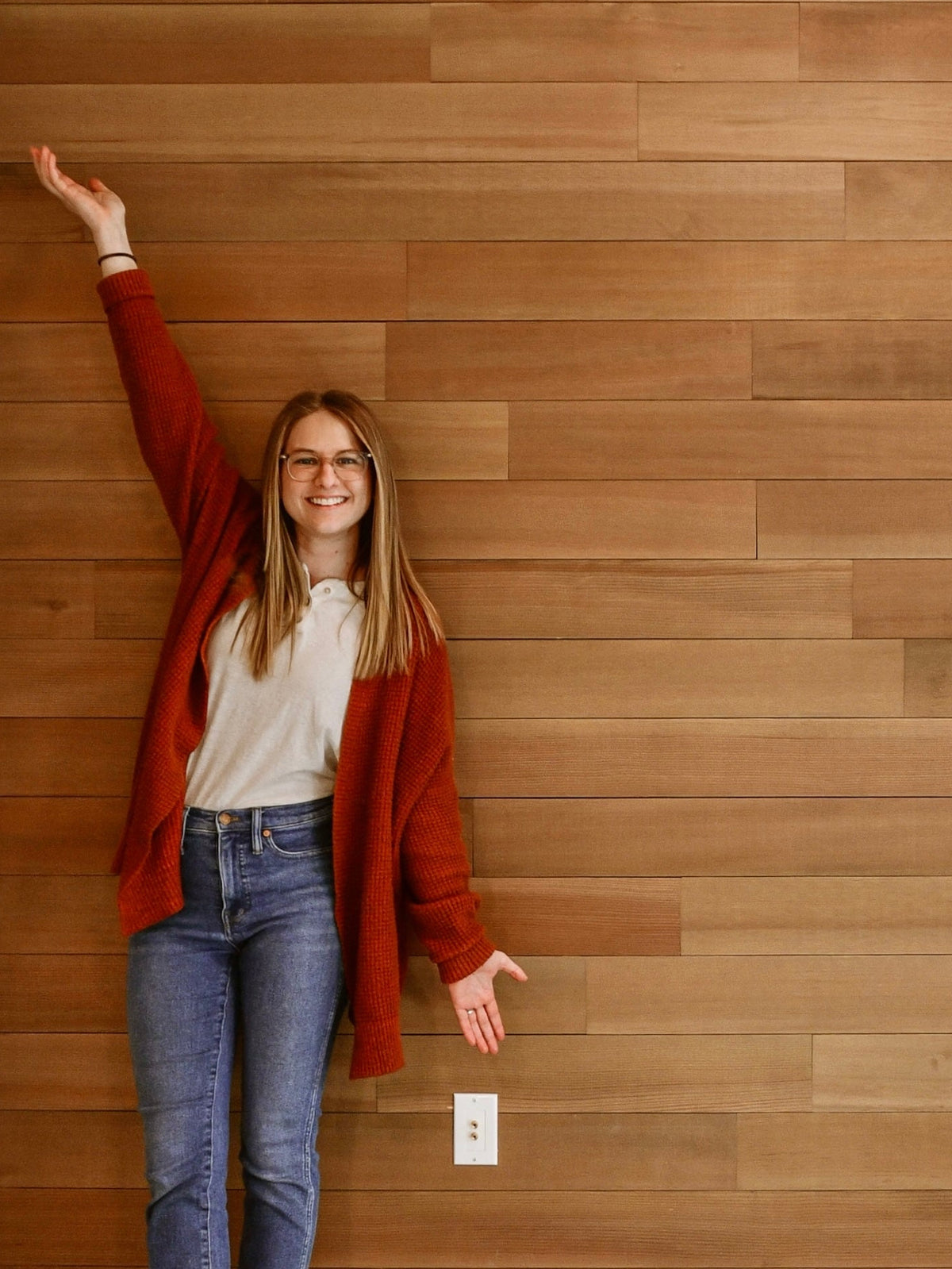 A woman stands in front of a wall made of horizontal wood planks, wearing a rust red cardigan, white top, blue jeans, and white sneakers, with one arm raised toward the wall.