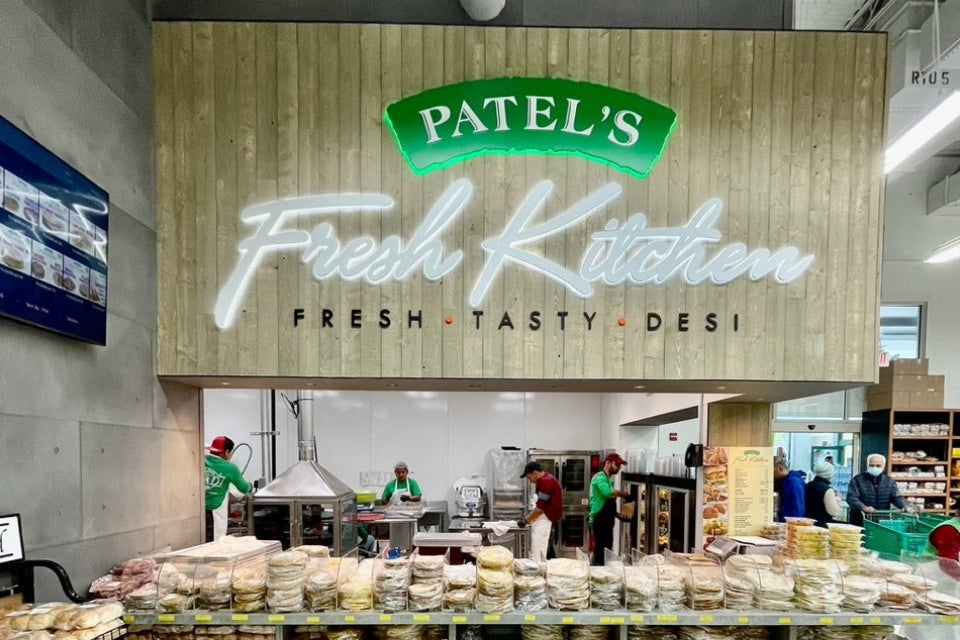 A large retail bakery display under a wooden sign reading 'PATEL'S Fresh Kitchen' with 'FRESH • TASTY • DESI' below. Shelves in front are stacked with assorted breads, pastries, and baked goods.